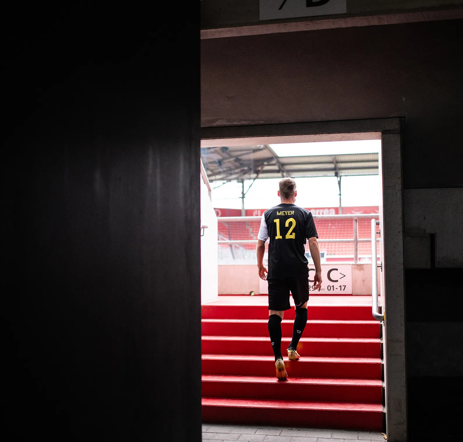 A soccer player in a black jersey with 'MIEYER 12' in yellow walks up the red stairs of a stadium.