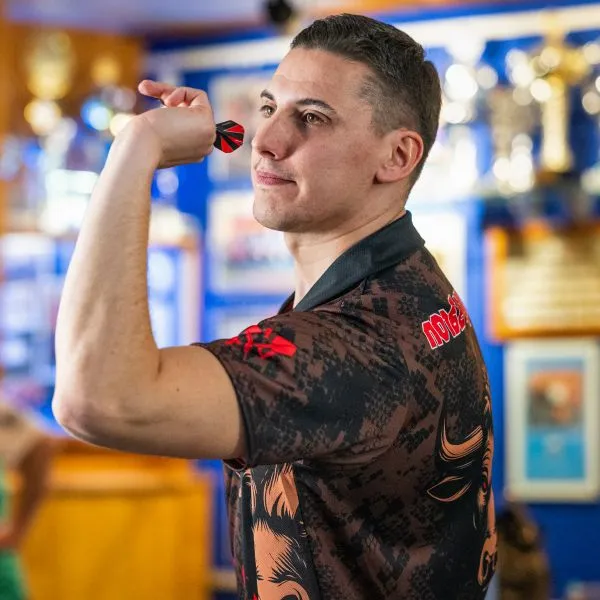A man in a patterned sports jersey with red accents holds a dart and aims, with trophies visible in the background.