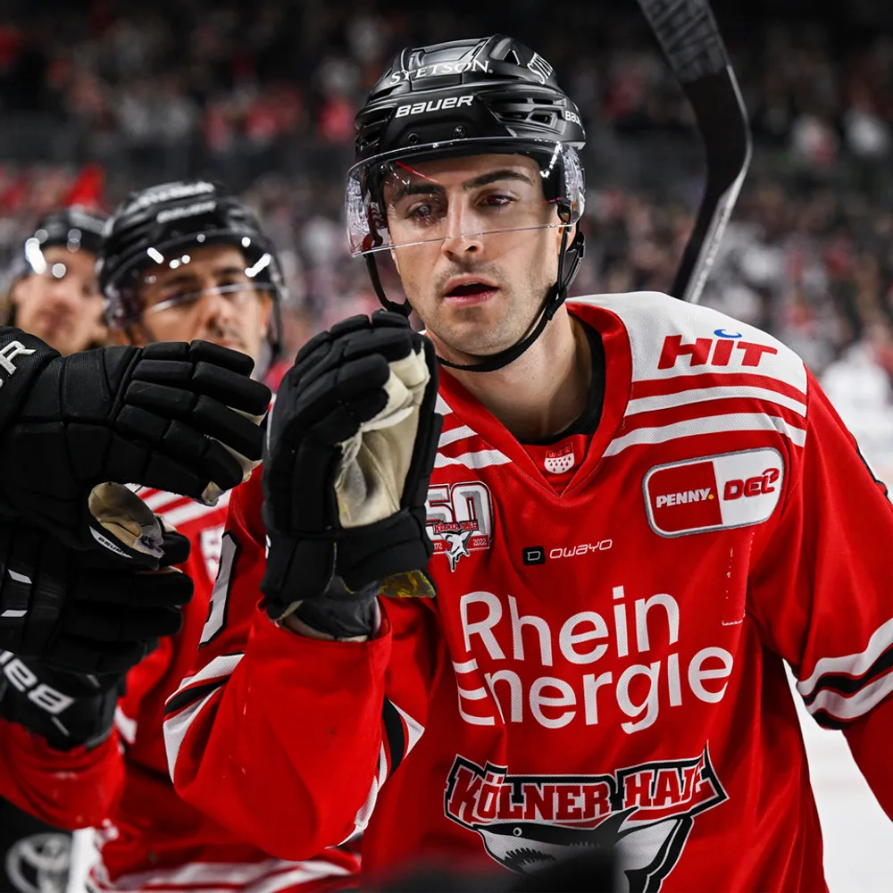 A hockey player in a red jersey with white stripes and logos, raising his hand in celebration.