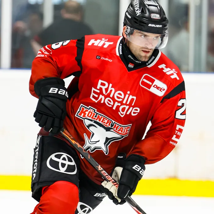 A hockey player in a red jersey with the Rhein Energie and Kölner Haie logo, standing on the ice.