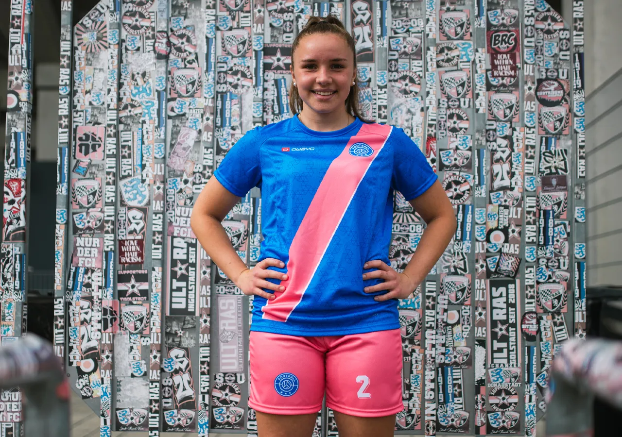 A girl wearing a blue soccer jersey with a pink diagonal stripe and pink shorts, smiling in front of a wall decorated with stickers.
