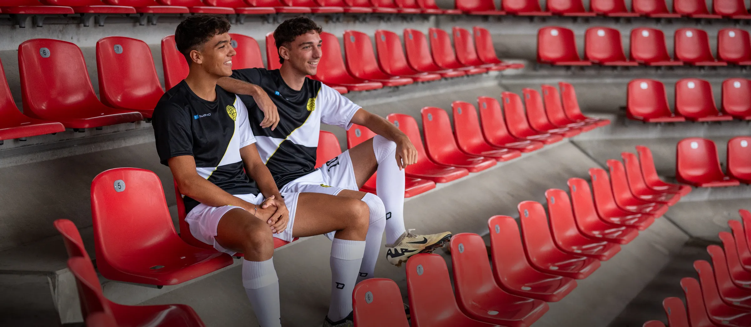 Two young men in sporty soccer jerseys are sitting casually on red stadium seats.