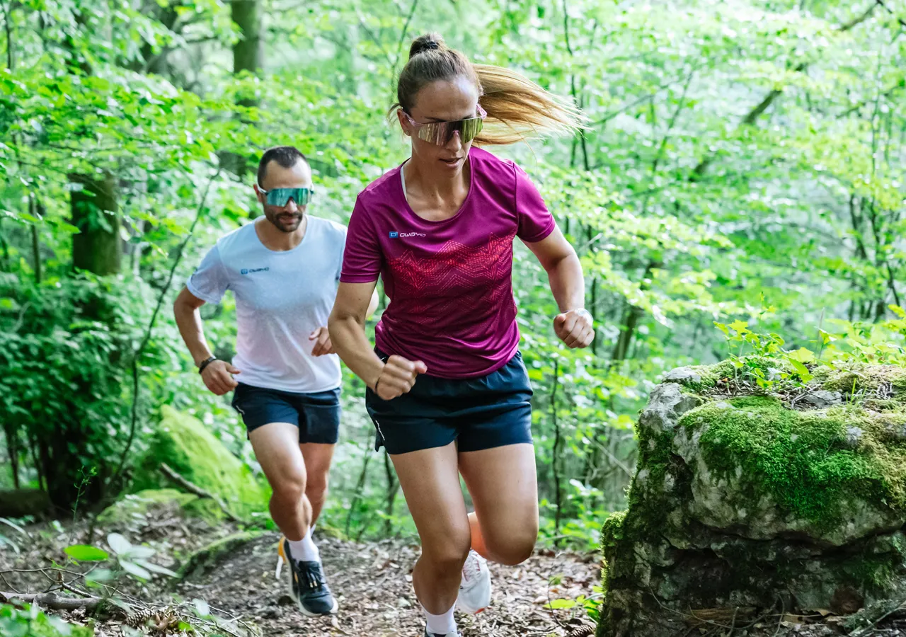 Two runners, a man in a white t-shirt and a woman in a red sports shirt, are running along a wooded path.