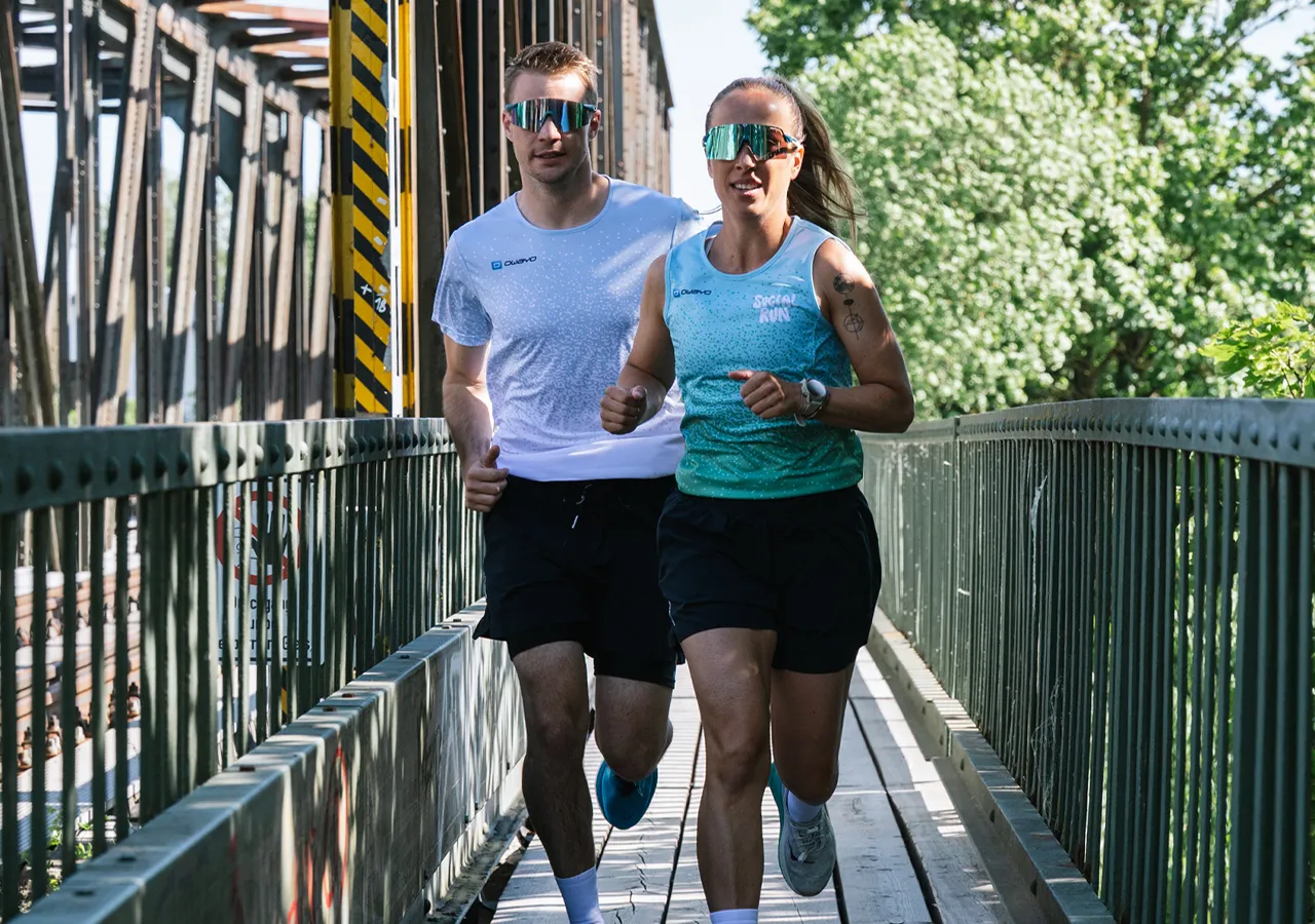 A man and a woman are jogging on a bridge, both wearing athletic clothing and sunglasses.