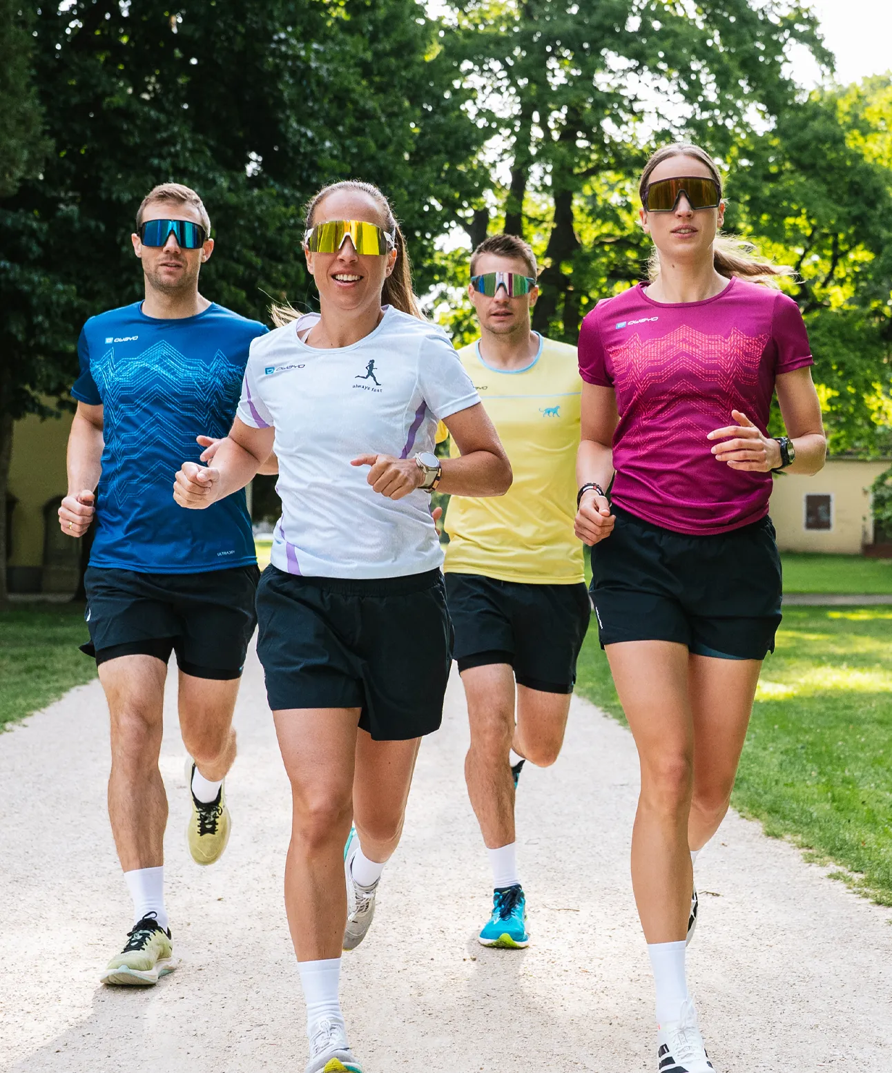 Four athletic individuals are running on a narrow path in a park, wearing colorful running shirts and sunglasses.
