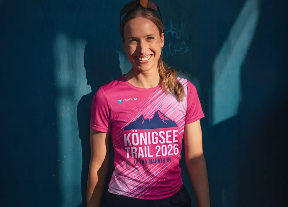A smiling woman is wearing a pink t-shirt with the print 'Königsee Trail 2026 Ultra Marathon' in front of a blue wall.