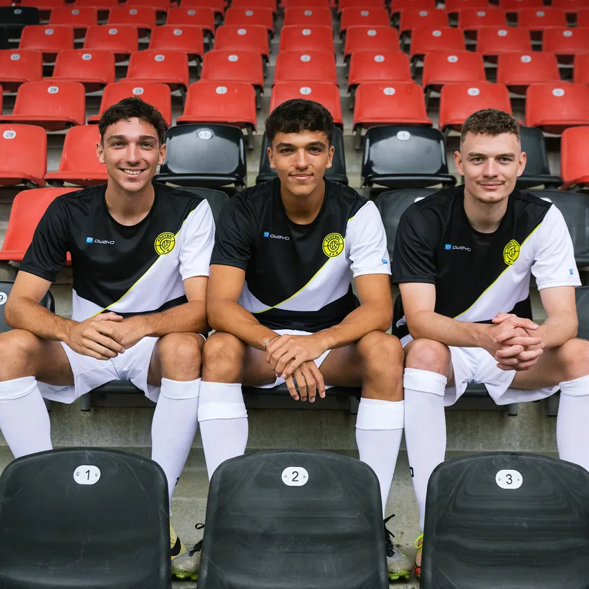 Three young men in owayo sportswear sit in a stadium on numbered seats in front of red bleachers.