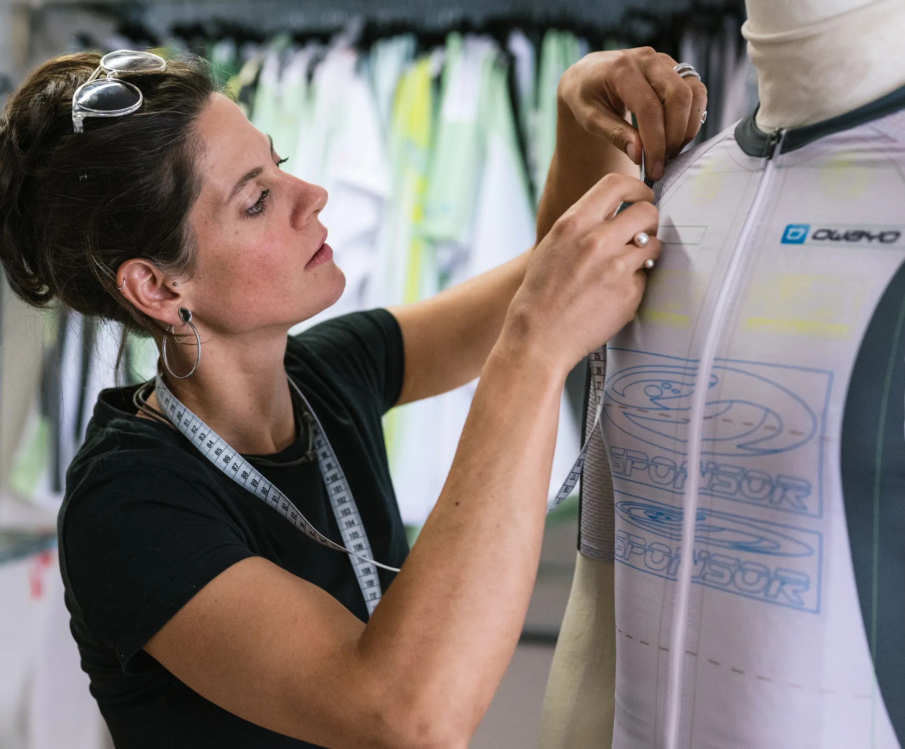 A woman measures a custom sports jersey on a mannequin, wearing a measuring tape around her neck.