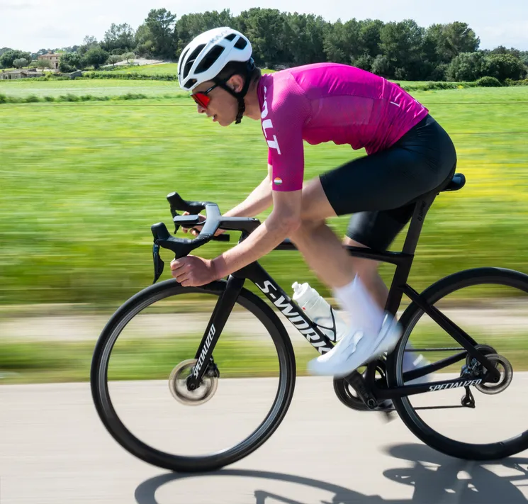 A cyclist in a pink jersey and black shorts rides on a road through a green landscape, with motion blur emphasizing speed.