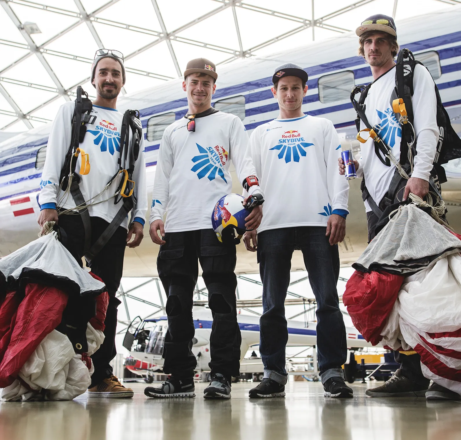 Four men in white long-sleeved sport shirts with the Red Bull Skydive Team logo stand in a hangar in front of an airplane.