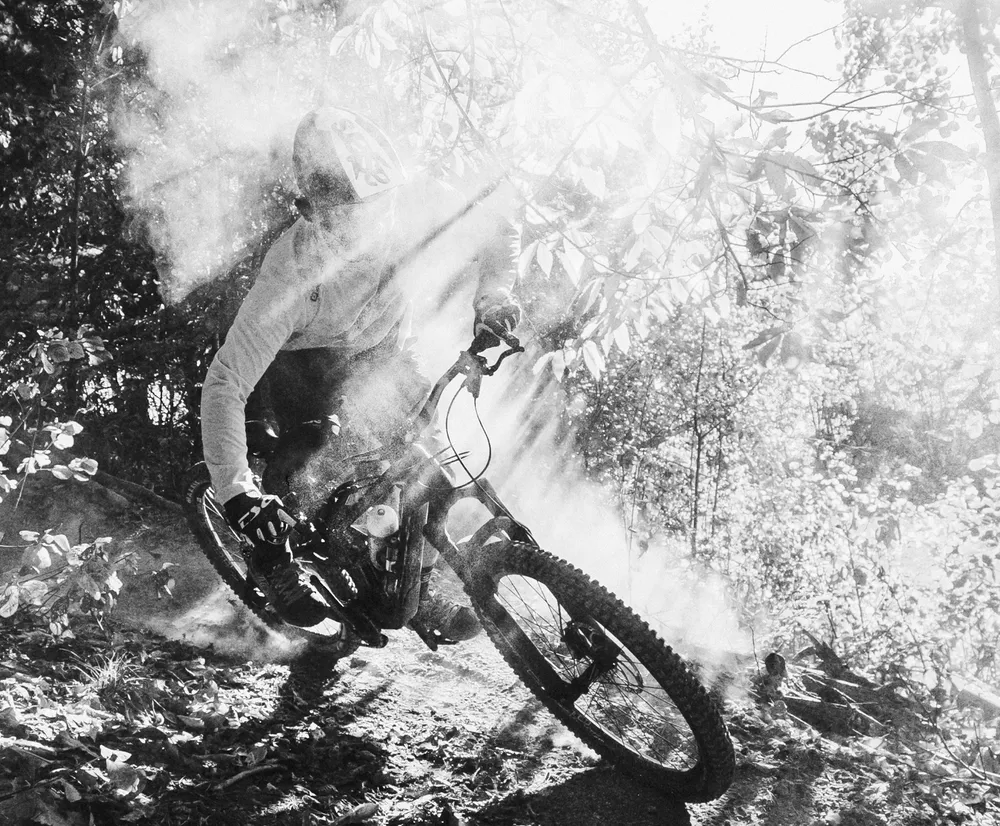 A mountain biker in motion on a forest trail, surrounded by dust and light rays, wearing a long-sleeved top from owayo.