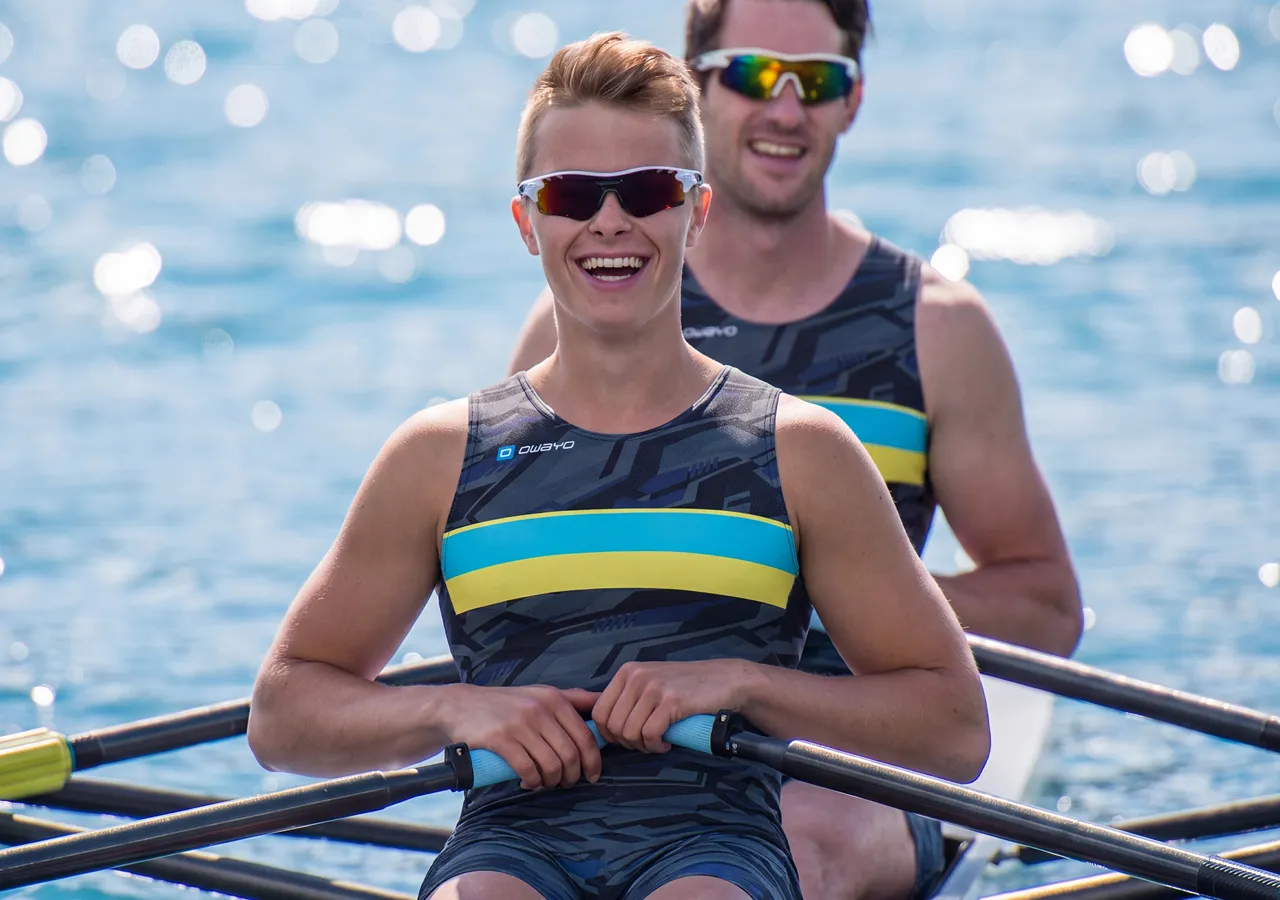 Two athletes in stylish sportswear sit in a rowing boat, smiling at the camera while paddling on shimmering water.