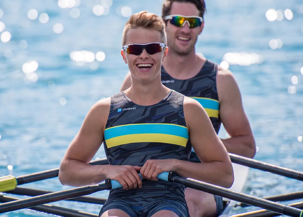 Two rowers in sporty patterned one-pieces with blue and yellow stripes, smiling on a body of water.