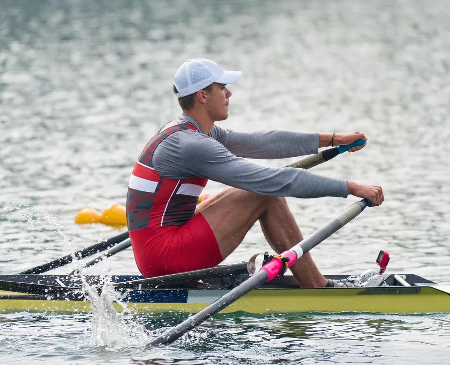A rower in a gray long-sleeve shirt and red shorts is rowing in a boat on calm water.