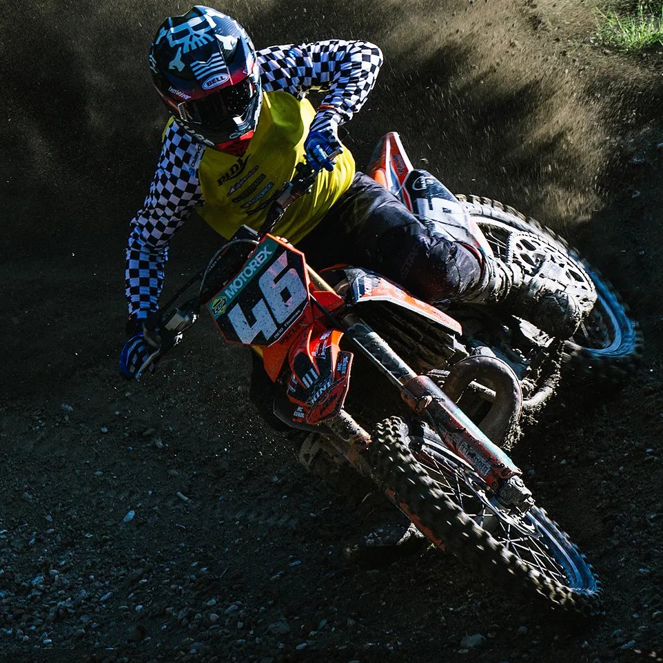 A motocross rider in a yellow jersey with black and white checkered sleeves and a helmet rides an orange motorcycle on a dusty track.