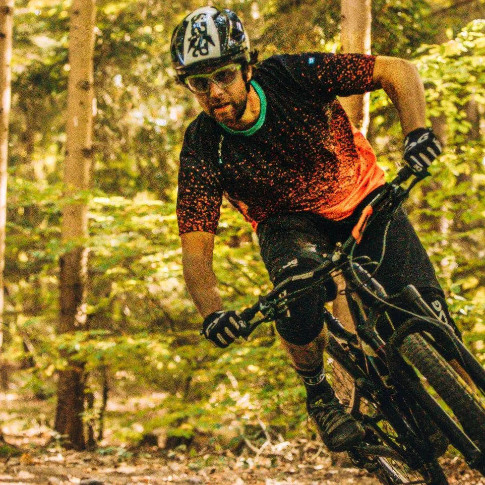 A mountain biker in a black, red-speckled jersey and black shorts rides through a forest.