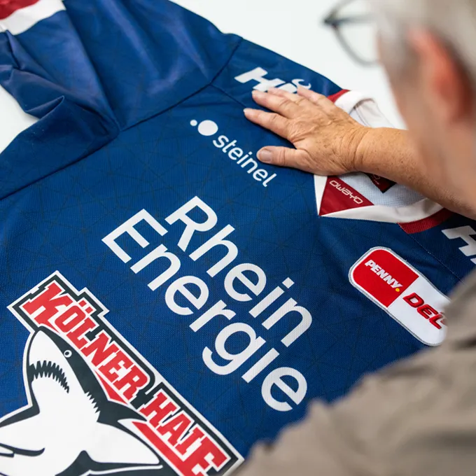 A person is inspecting a blue customized sports jersey from owayo featuring various sponsor logos, including 'Rhein Energie' and 'Kölner Haie'.