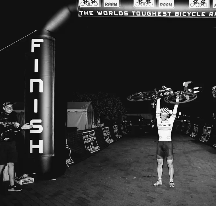 A cyclist celebrates crossing the finish line of the 'Race Across America' with his bike raised above his head, as a man applauds beside him.