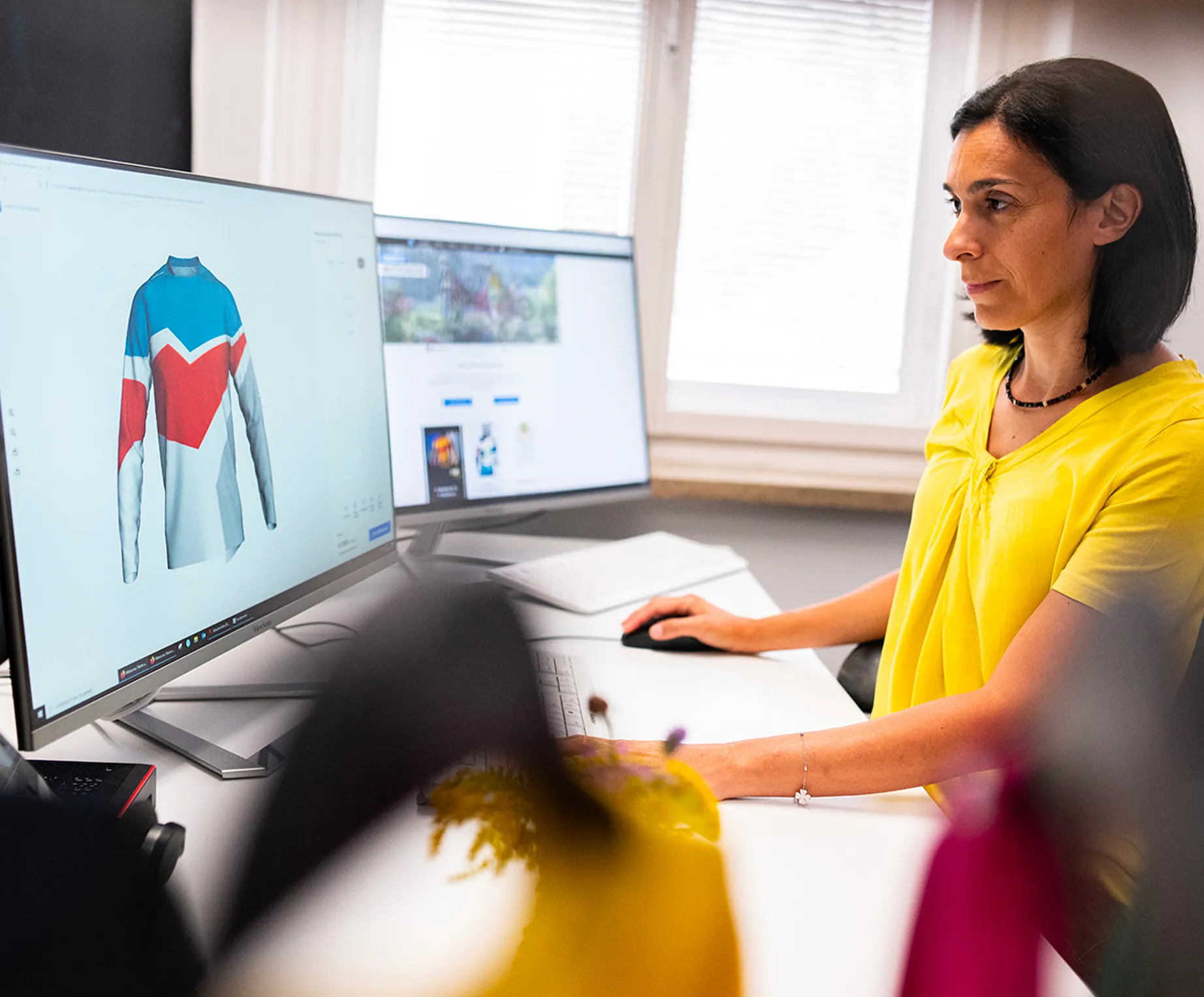 A woman in a yellow top sits at a desk and designs a customized sports jersey with blue, red, and white panels on a computer screen.